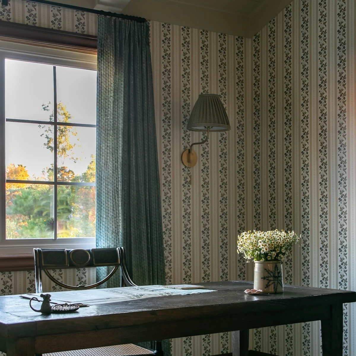 an image of a desk with wallpaper samples on it. There is wallpaper on the walls, and curtains on the windows. 