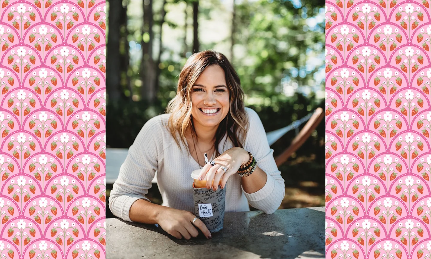 a woman sits at a picnic table, smiling and holding an iced coffee in a fabric sleeve. 