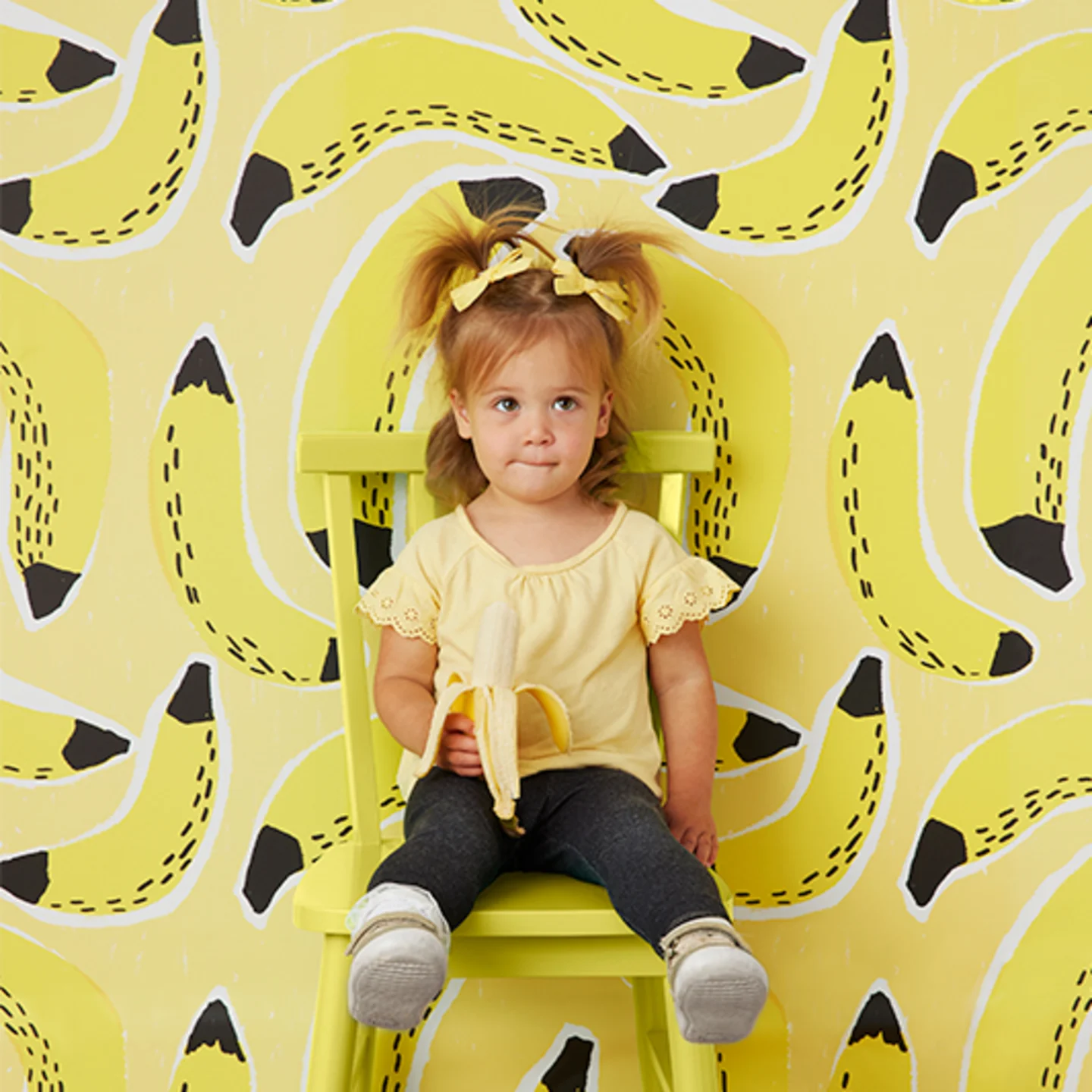 Little girl eating a banana next to banana wallpaper.