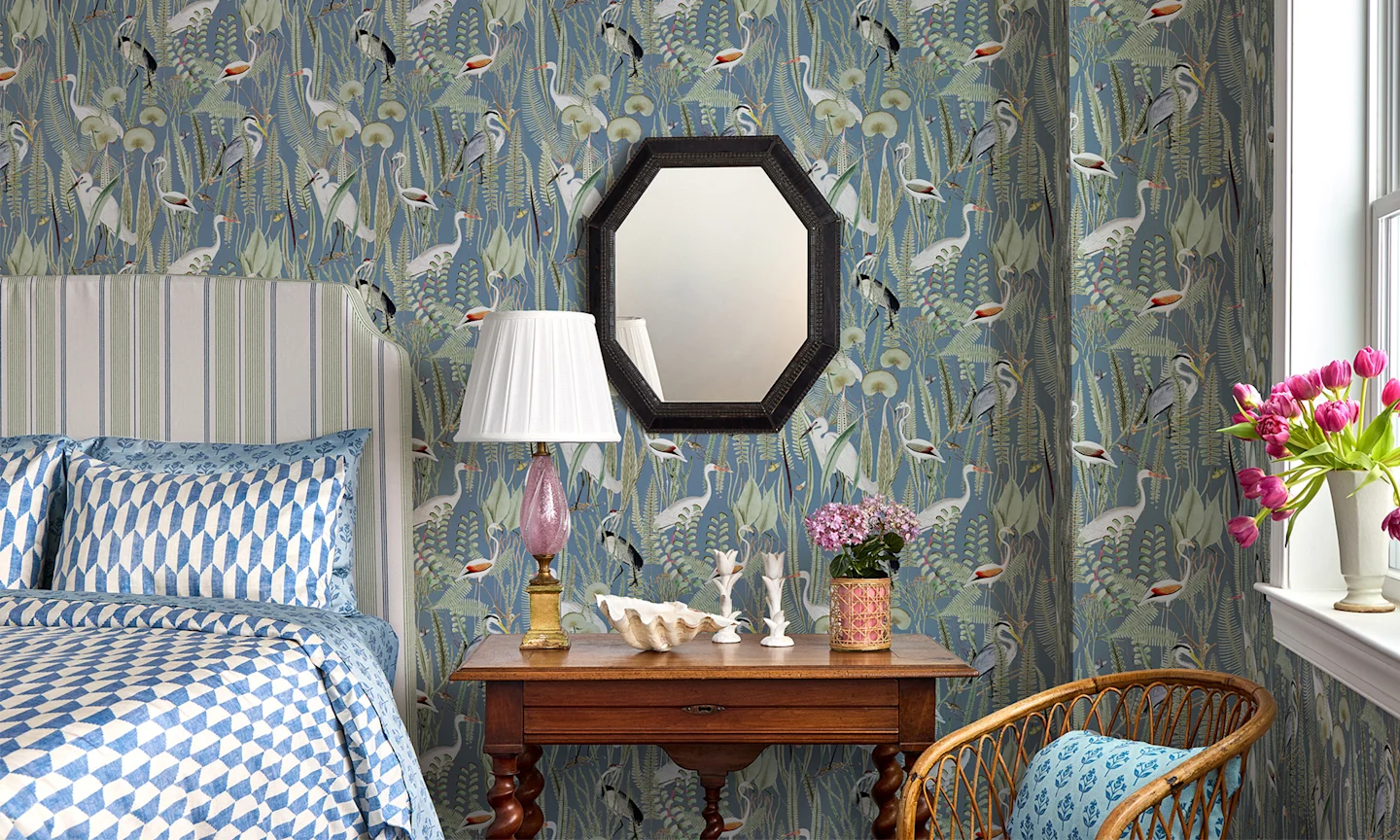 Bedroom with coastal heron wallpaper a striped headboard and geometric blue and white bedding.