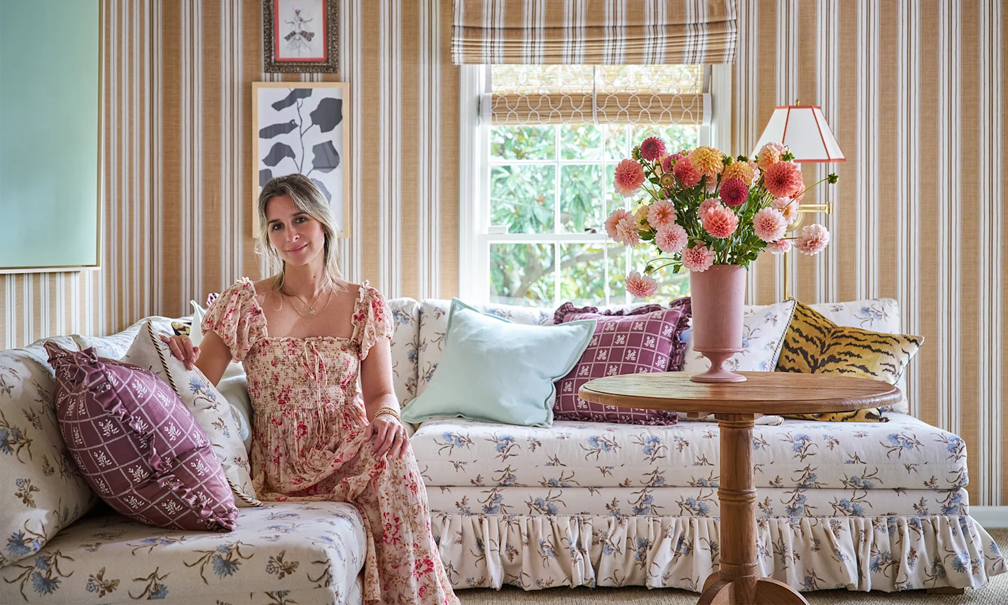 Whiteney McGregor in a living area she designed with striped wallpaper on the walls and a couch upholstered in floral fabric.